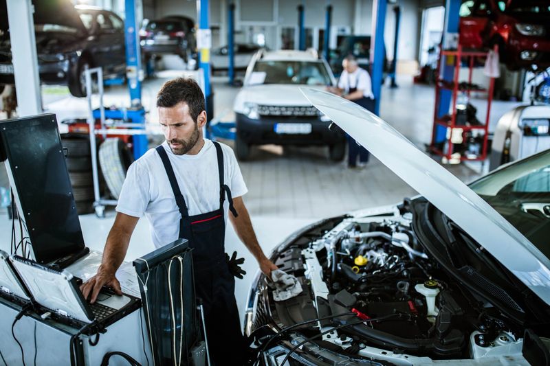 Young mechanic using computer while working on car diagnostics in a repair shop.