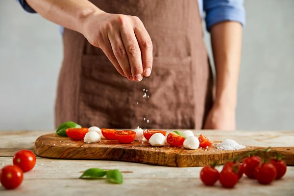Person sprinkling salt over tomatoes and mozzarella on a wooden board.