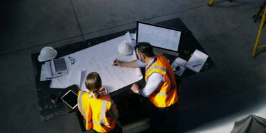Two construction workers review blueprints on a table in a warehouse.