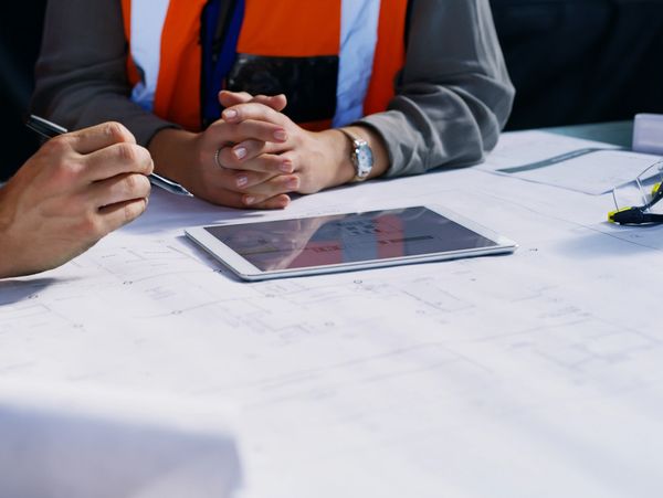Two workers in orange vests review blueprints with a tablet and notes on a table.