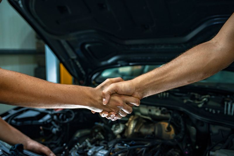 Caucasian young mechanic shaking hands with a mature male customer in front of a car. Photo of a  man giving an handshake in an industrial facility. Man greets auto master. Concept - car service. Handshake.