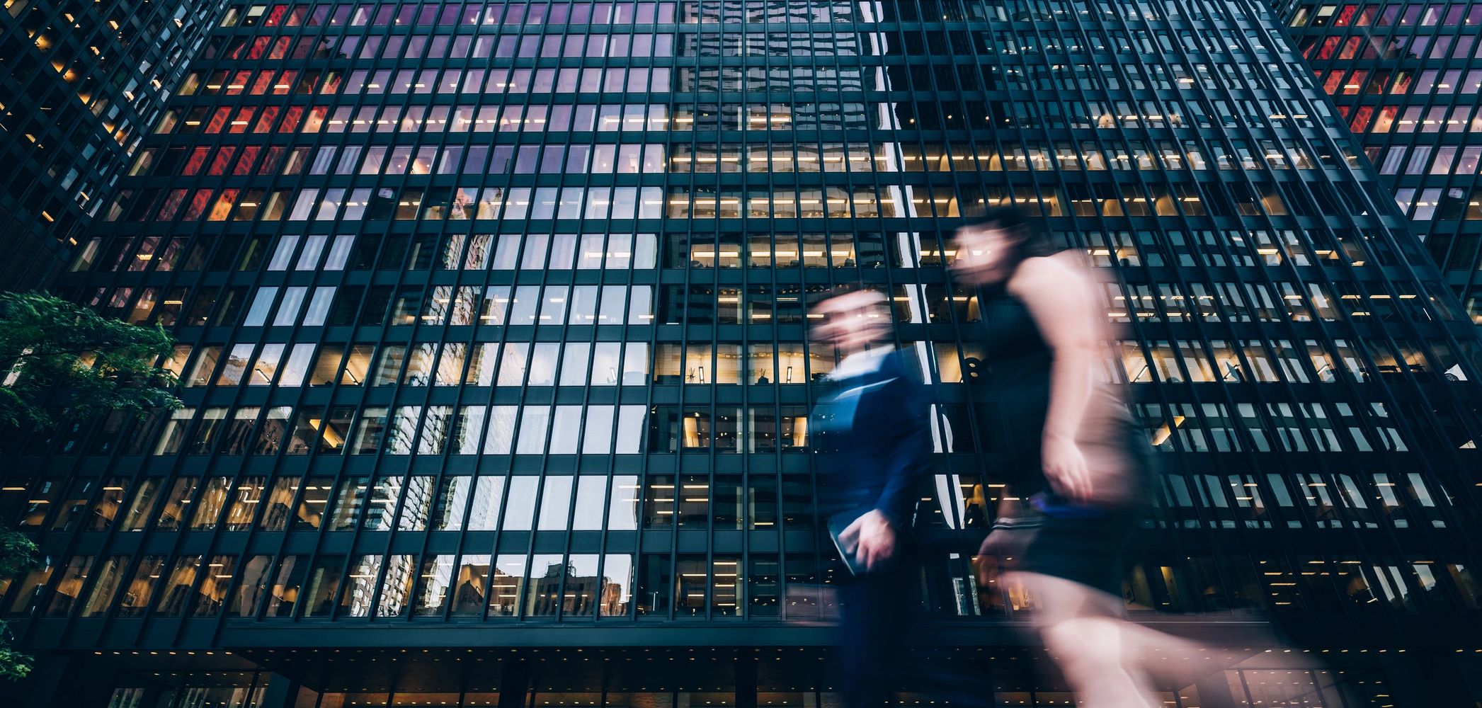 Blurred figures walking past a reflective glass office building.