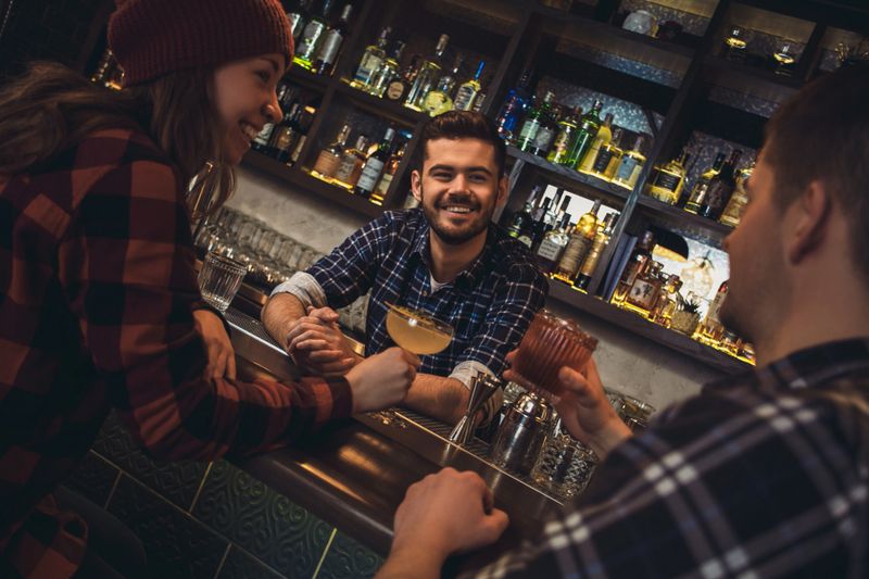 Young barman standing at bar counter with visitors drinking alcohol talking smiling joyful