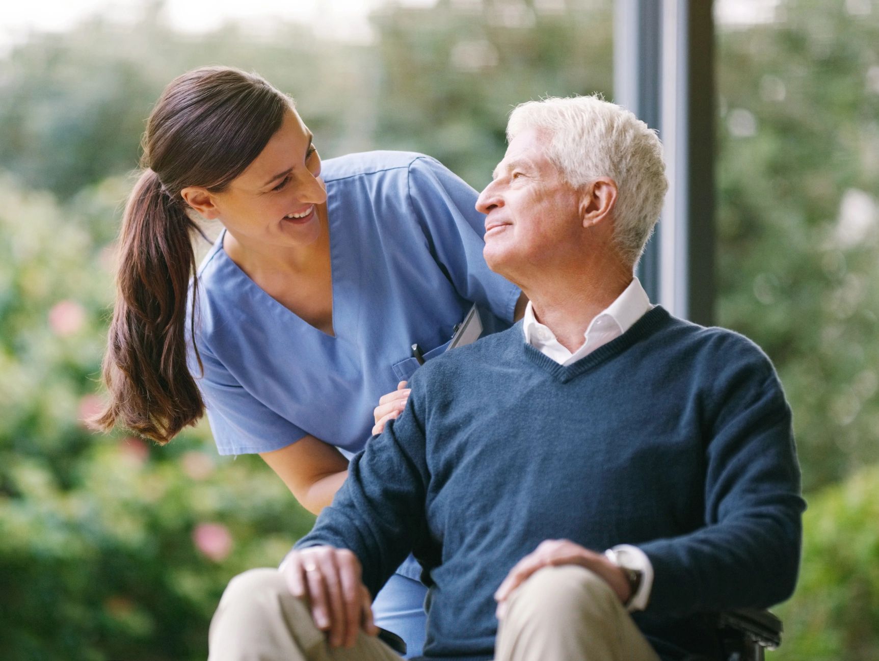 Caregiver smiling with an older adult in a wheelchair outdoors, compassionate assisted living care.