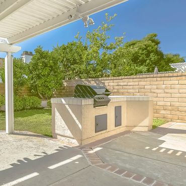 Outdoor built-in barbecue grill under a white pergola in a sunny backyard.