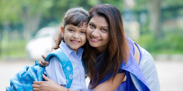 A mother happily hugging her smiling daughter with a school backpack outdoors.