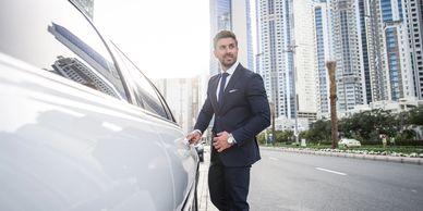 A man in a suit opens a car door in a city with tall buildings.