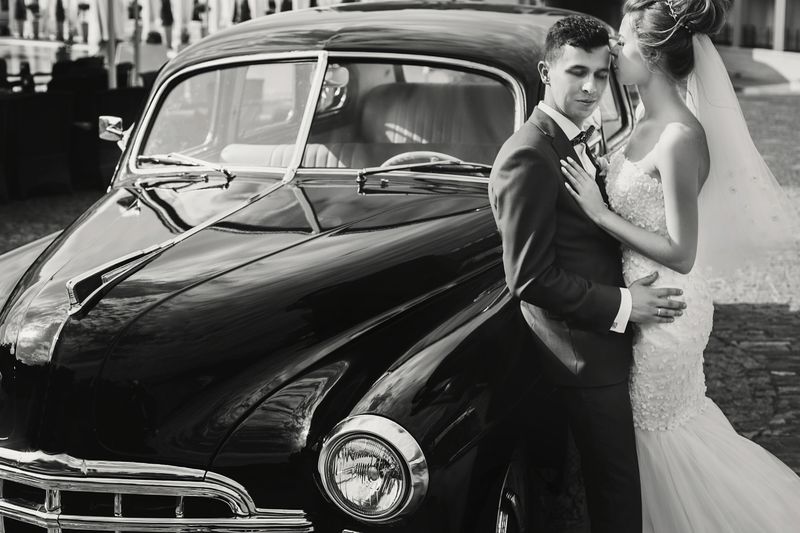Stylish happy bride and groom kissing at old black retro car. Gorgeous wedding couple of newlyweds embracing after wedding ceremony. Romantic moment