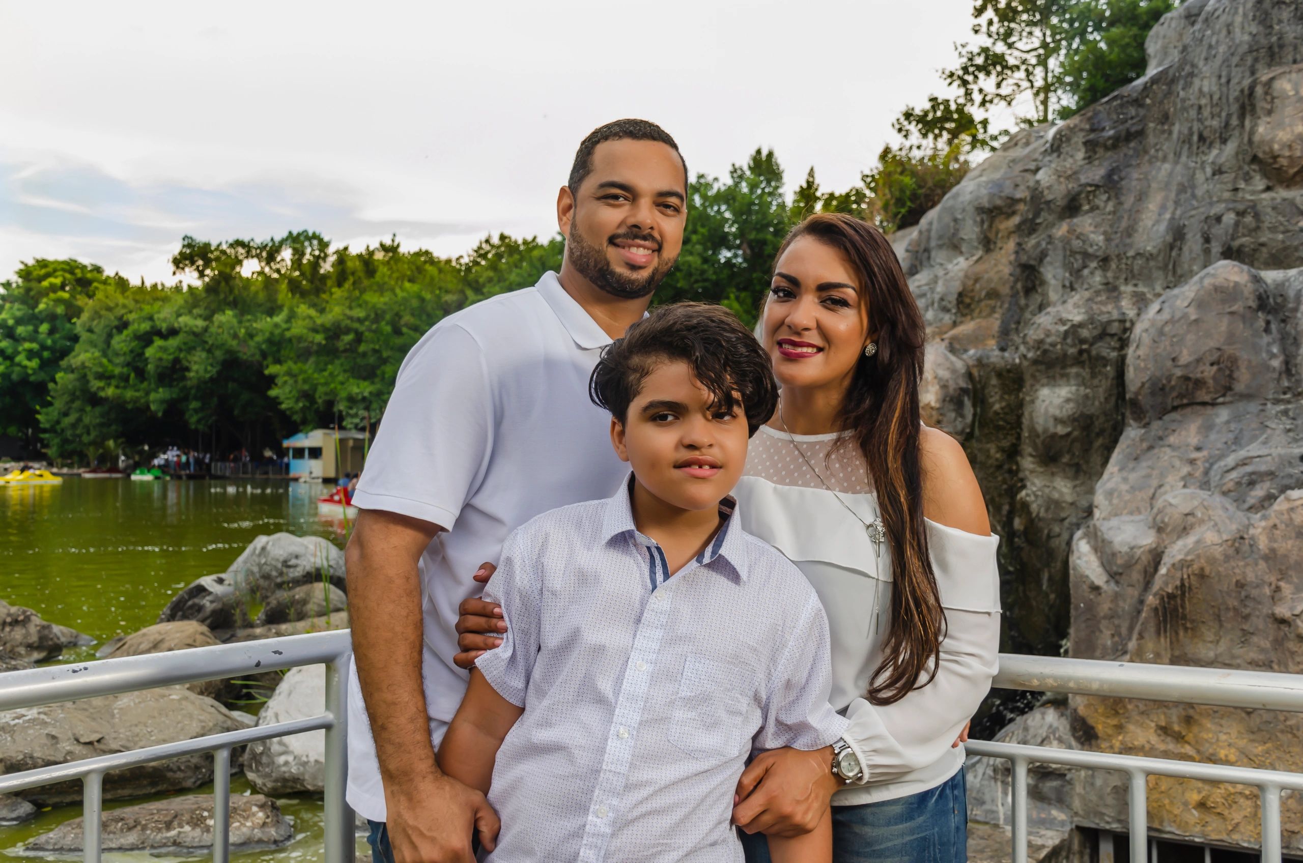A happy family of three posing outdoors near a rocky lake.