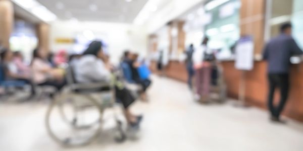 Blurred image of people waiting in a medical facility, including a person in a wheelchair.