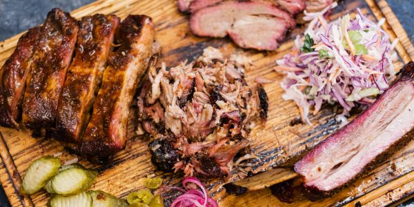 A wooden platter with various smoked meats, pickles, and coleslaw.