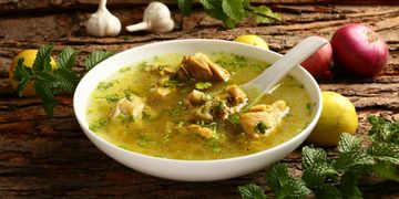 A bowl of chicken soup garnished with herbs, surrounded by garlic, onions, lemons, and fresh mint.