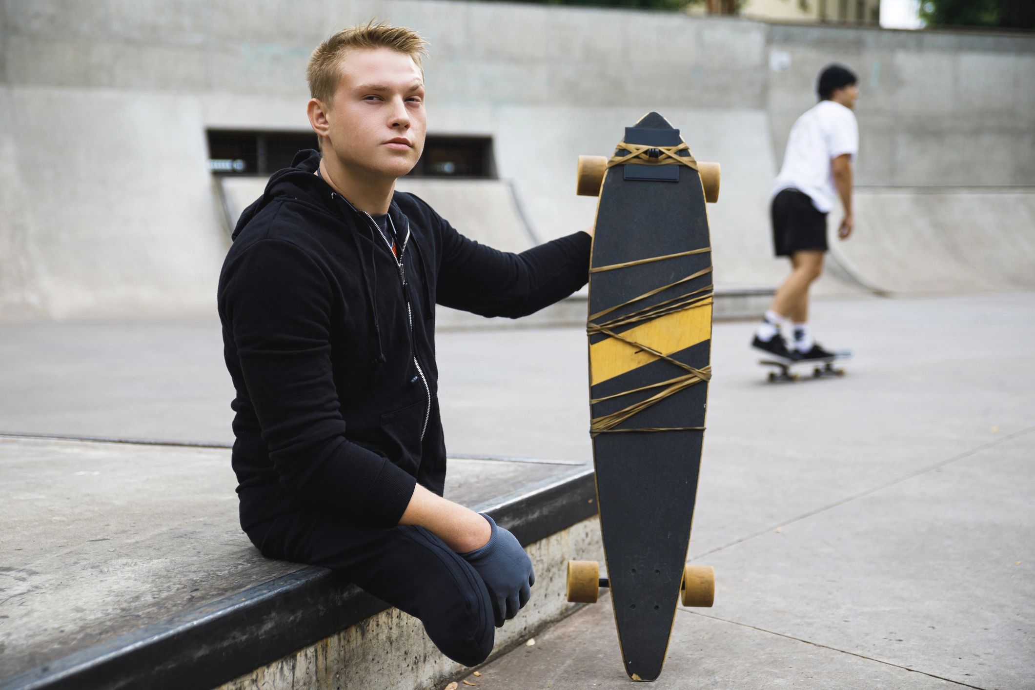 Young man sitting with a longboard at a skatepark, another person skateboarding in the background.