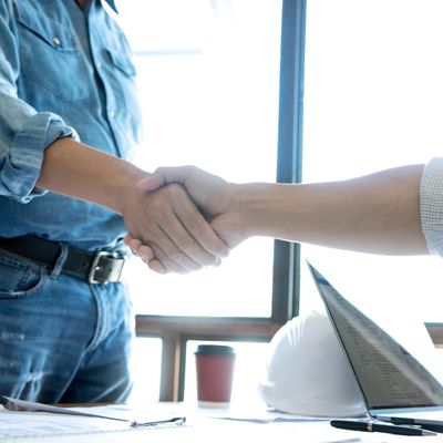 Two people shaking hands over a desk with construction plans and a laptop.