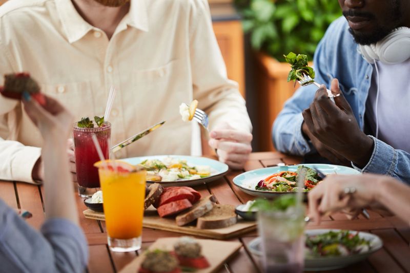 Close-up of young people sitting at the table eating vegetable salad and drinking juice during lunch in cafe