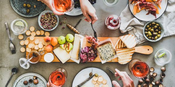 A charcuterie board with cheese, meats, fruits, and crackers surrounded by drinks and hands.