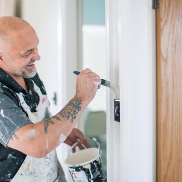 A smiling man with tattoos painting a white door indoors.