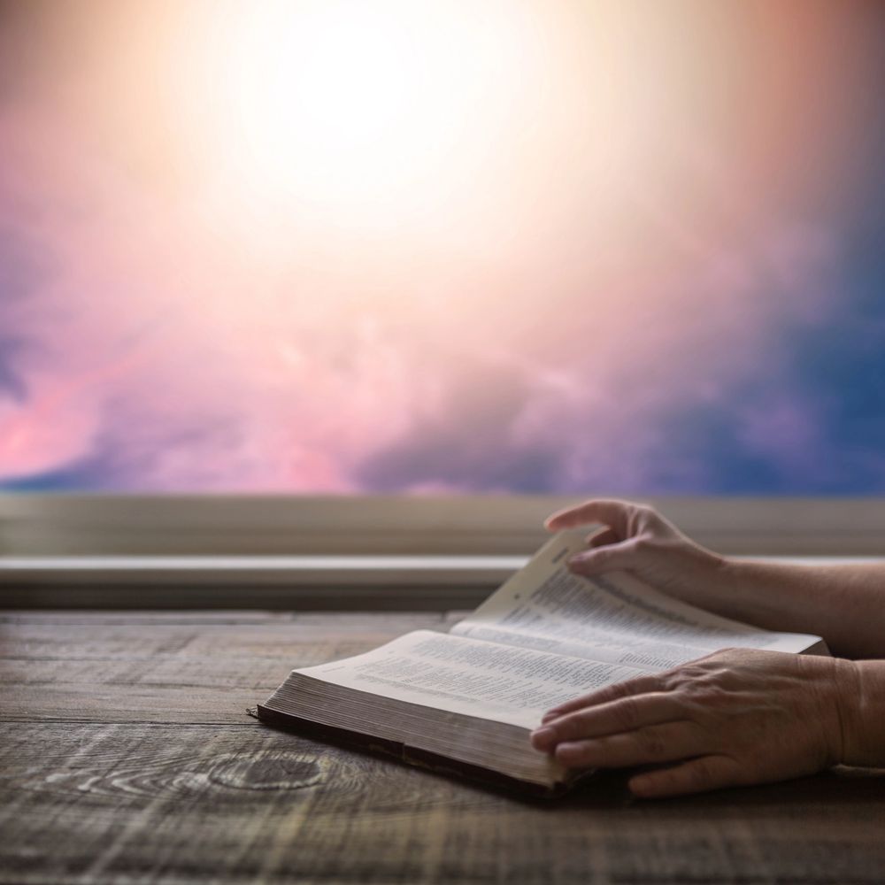 Hands gently turning pages of an open book on a wooden table with a glowing sky background.