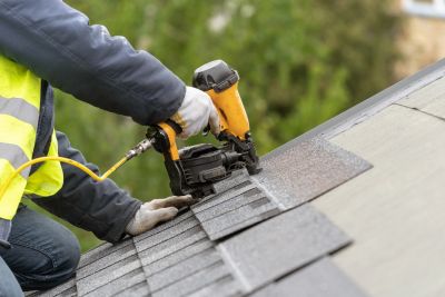 Worker installing asphalt shingles on roof with nail gun.