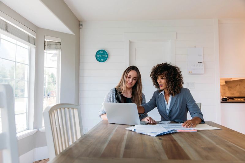 A female financial advisor sits with a woman at her dining room table with laptop and financial reports helping her monthly budget and investments. She is showing her client how to read a financial report as she works on her computer to do monthly finances, pay taxes and save money for the future.