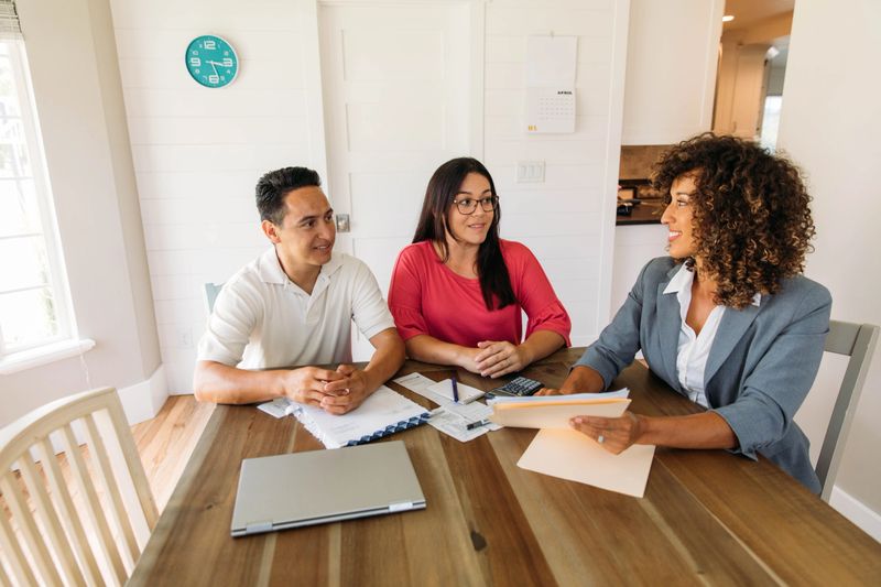 A female financial advisor sits with a millennial age married couple at a dining room table with laptop and financial reports helping with their monthly budget and investments. She is showing her clients how to read financial reports and monthly finances, pay taxes and save money for the future.
