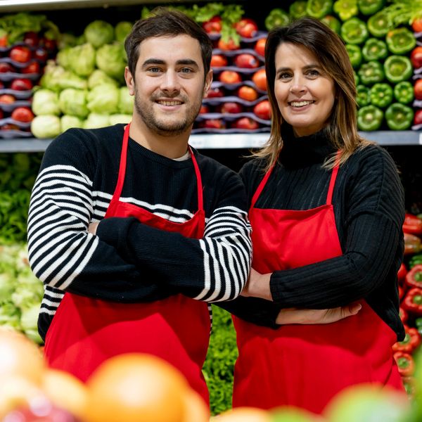 Two smiling grocery workers in red aprons stand confidently in front of fresh produce.