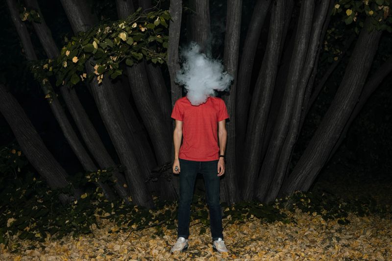 Young Man With Smoke Covering Face Standing Against Tree Trunk In Forest