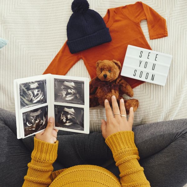 Pregnant woman holding ultrasound images with baby clothes and teddy bear nearby.