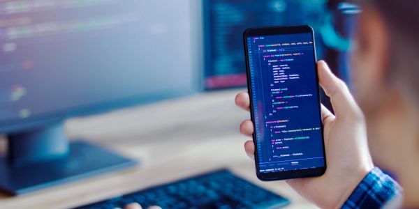 Person coding on a smartphone with a computer in the background.