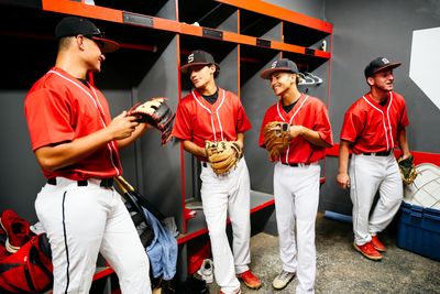 Four young baseball players in red uniforms chatting in a locker room.