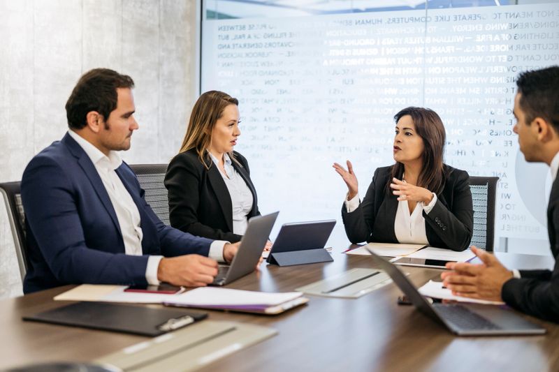Mature Hispanic businesswoman sitting at conference table with project team and sharing development updates.