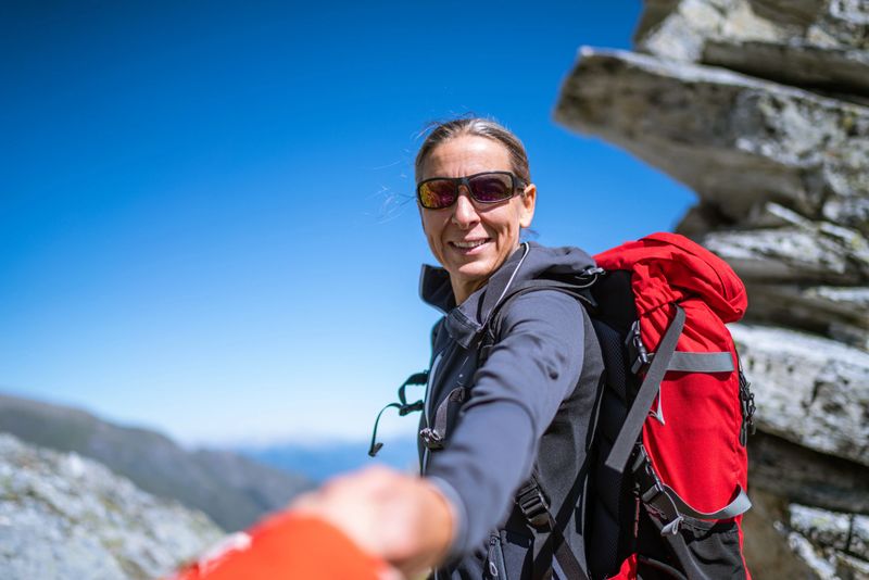 happy smiling hiking mature woman 45 years old with backpack and sunglasses high above timber line in mountains on sunny day looking back over her shoulder giving inviting supporting hand to follow her