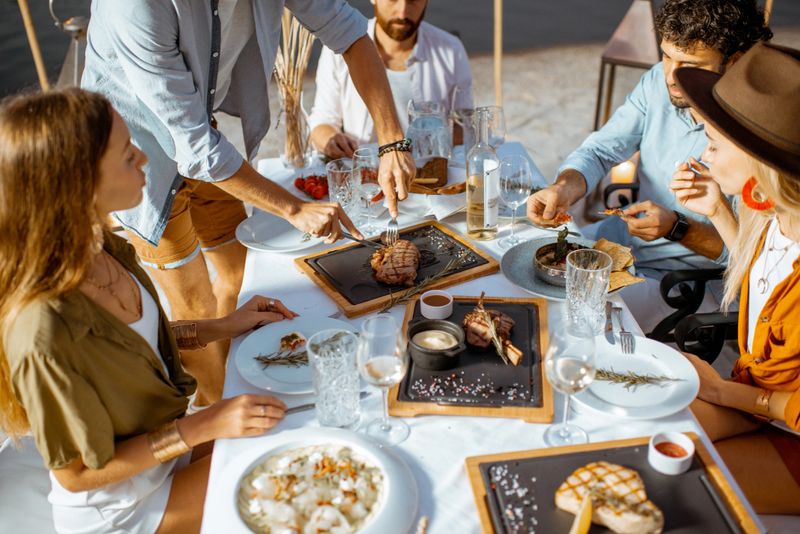 Group of friends having a festive dinner at the beautifully decorated table with delicious meals on the beach at dusk