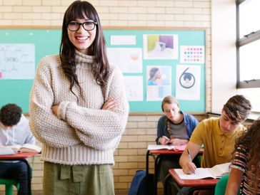 Smiling teacher stands confidently in front of focused students in a classroom.