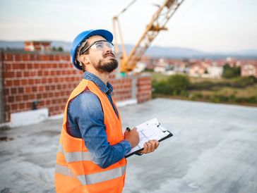 Construction worker in safety gear inspecting site with clipboard.