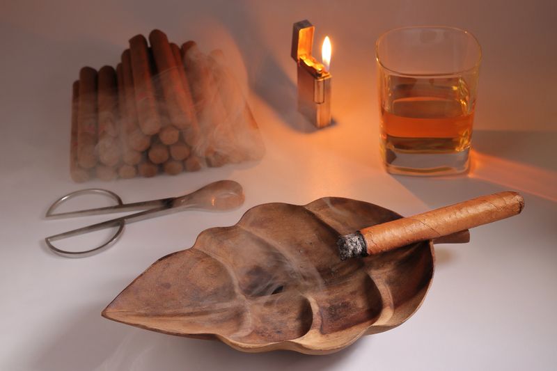 HAVANA CIGAR IN A WOODEN ASHTRAY NEXT TO A CIGAR CUTTER AND A LIT LIGHTER