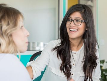 A woman with glasses smiling warmly while talking to another person.