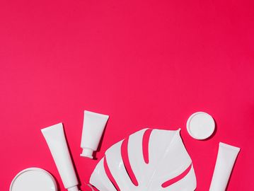 White cosmetic tubes and jars arranged with a white leaf on a bright pink background.