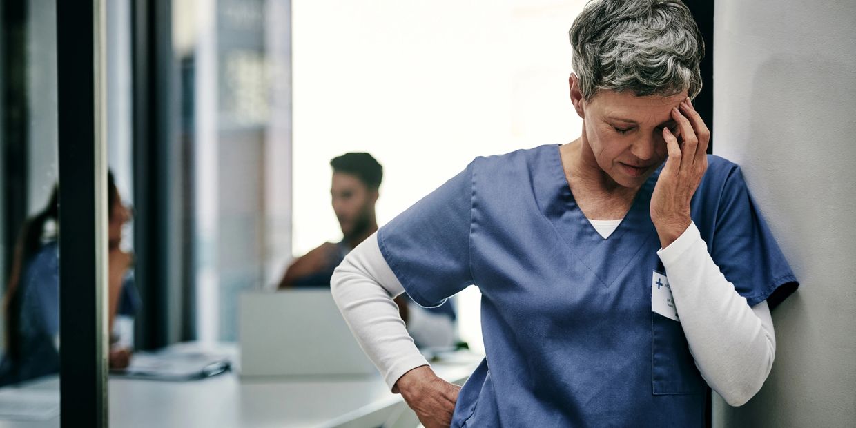 A nurse in scrubs looking stressed and leaning against a wall.