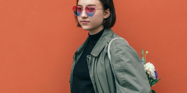 Young woman with heart-shaped sunglasses poses against an orange wall.