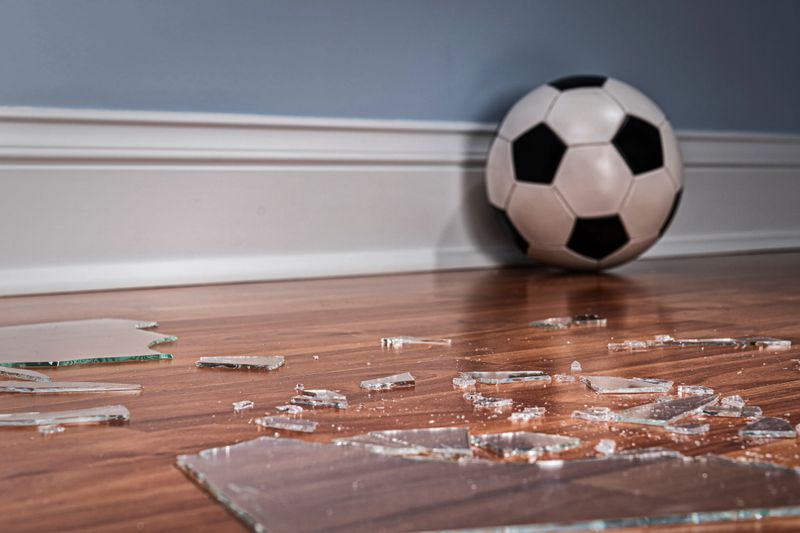 Pieces of glass from a broken window pane on the wood floor inside a home with a soccer ball in in the background.