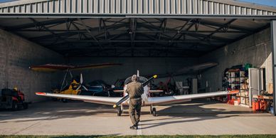 A person walks toward a small airplane inside a hangar on a sunny day.