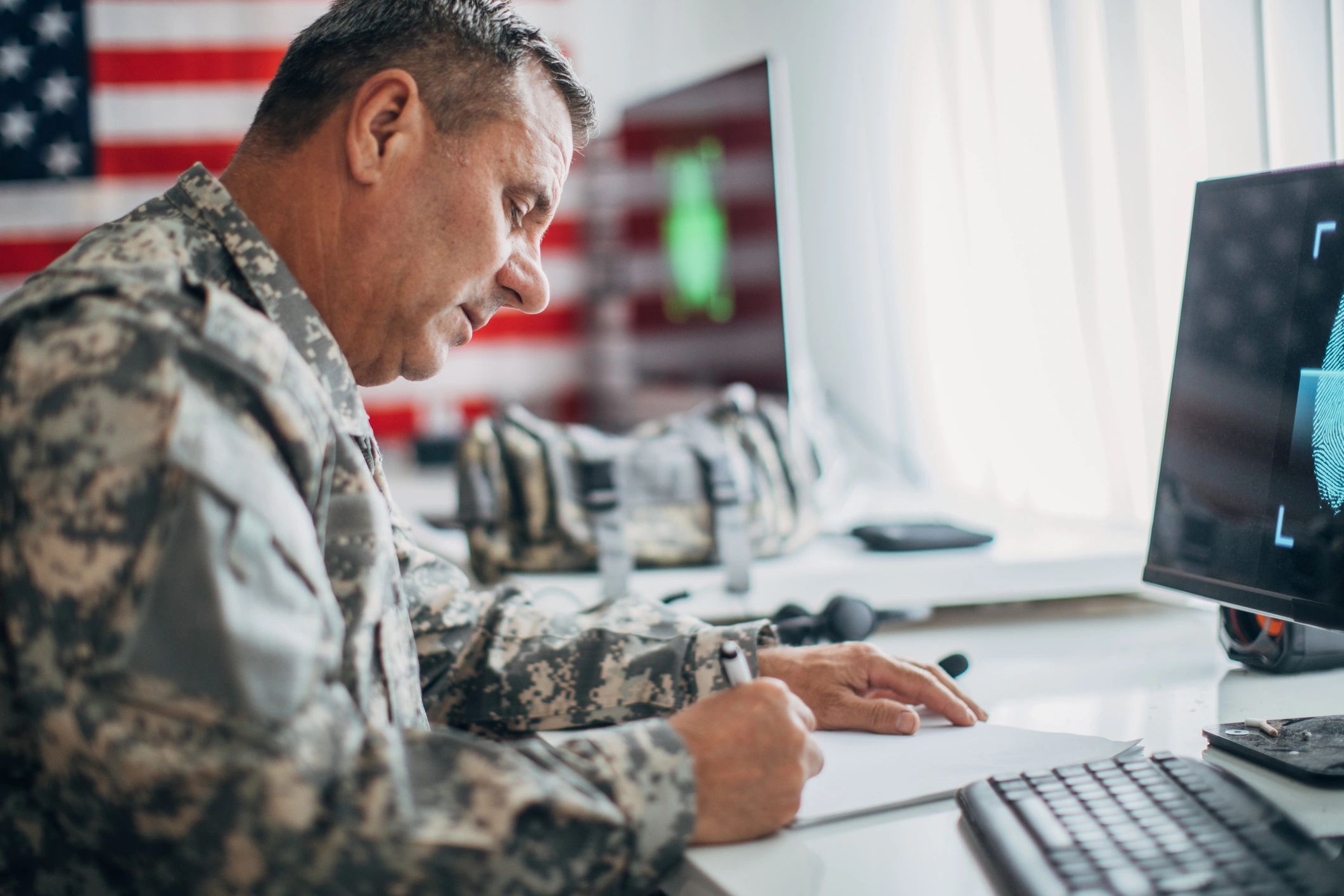 Military officer working at a desk with an American flag in the background.