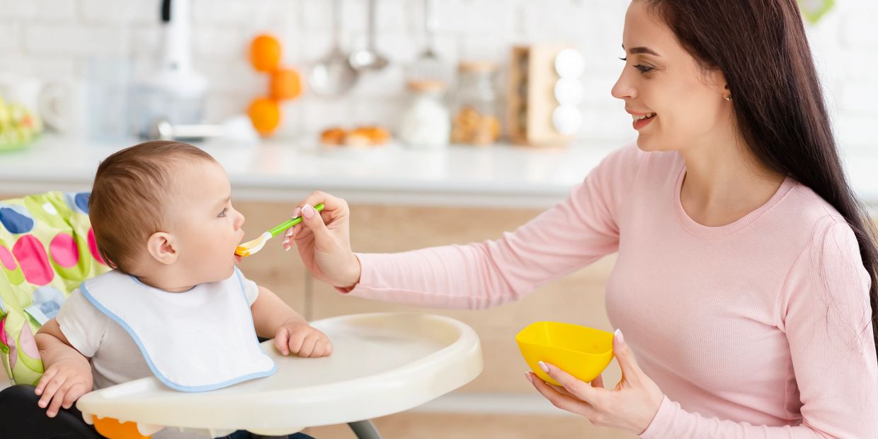 A mother feeding her baby sitting in a high chair in the kitchen.