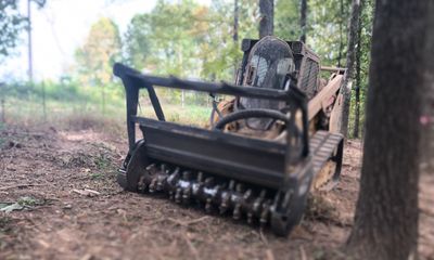 A forestry mulcher machine parked on a dirt ground in a wooded area.