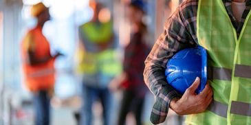 Construction worker holding a blue helmet with colleagues in the background.