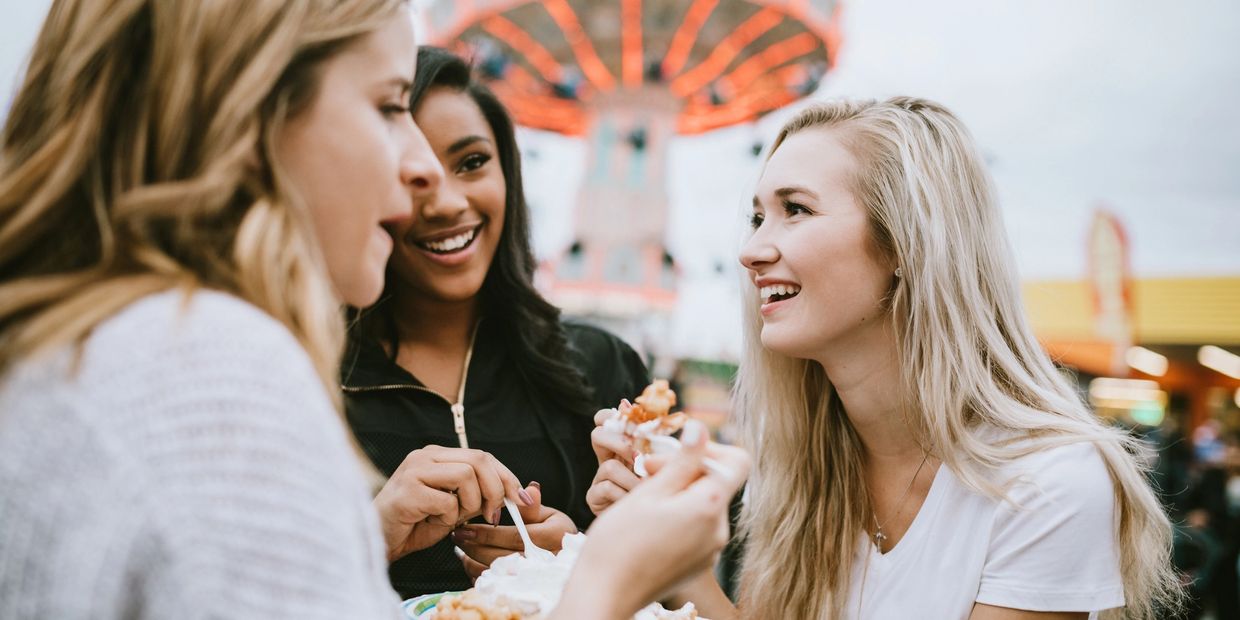 Three friends enjoying food and laughter at an amusement park.