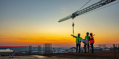 Three construction workers discuss plans at a building site during sunset.