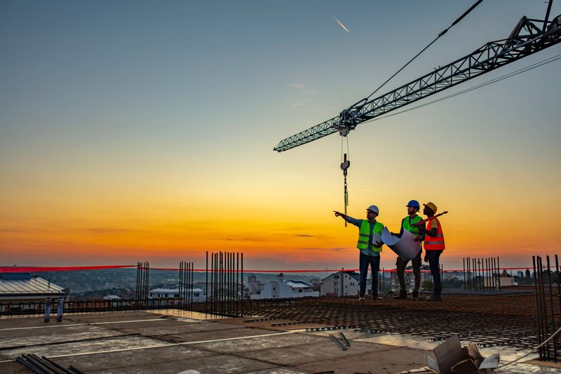Three Multi-Ethnic construction workers in uniform standing at construction site with crane in background, discussing building plans while holding blueprint at sunset under the tower crane.