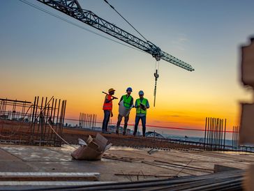Three construction workers in safety gear discussing at sunset on site.